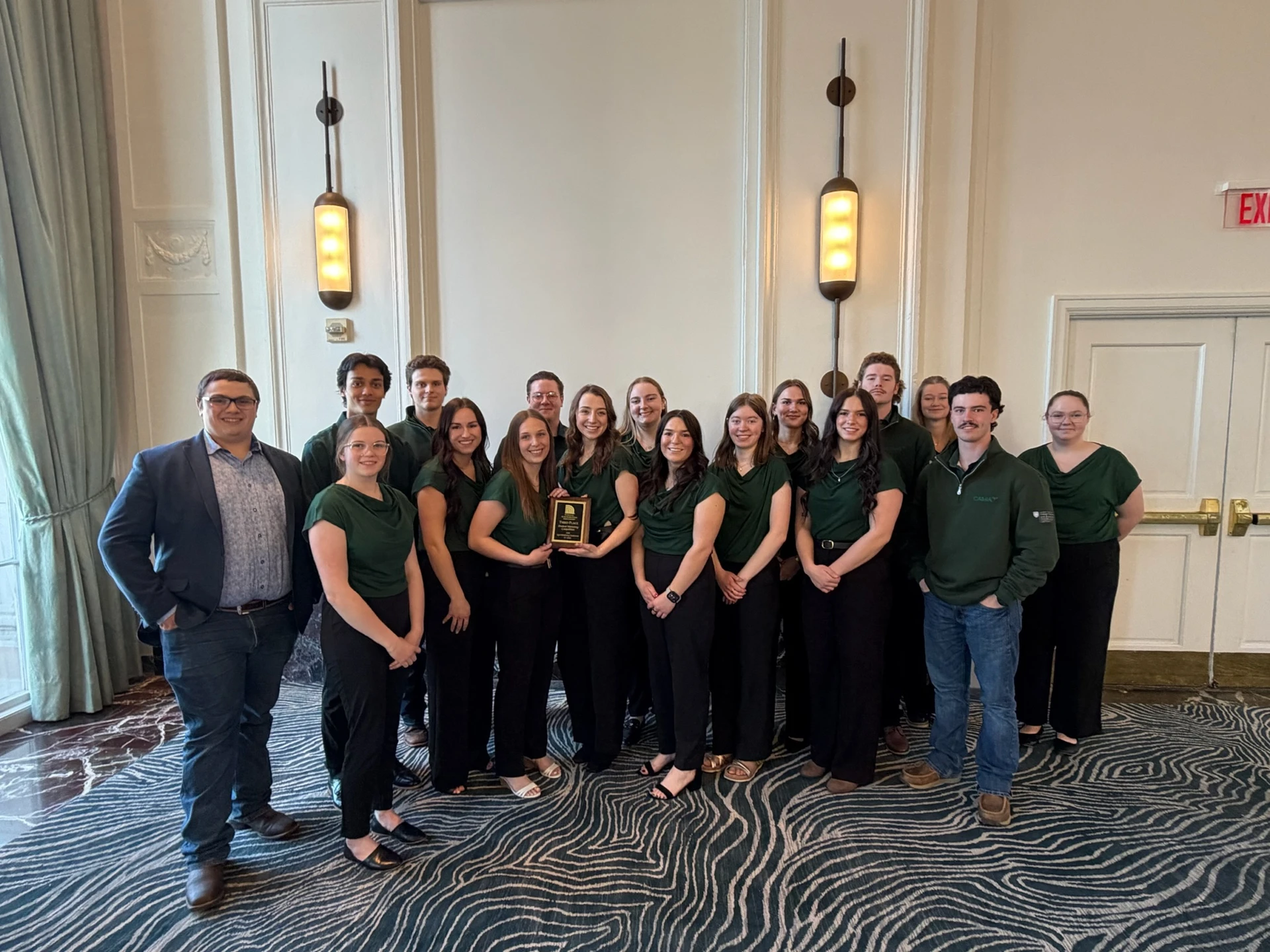 A group of fifteen young adults, most wearing dark green shirts and black pants, pose for a photo in a formal room. One woman in the center holds a small plaque.