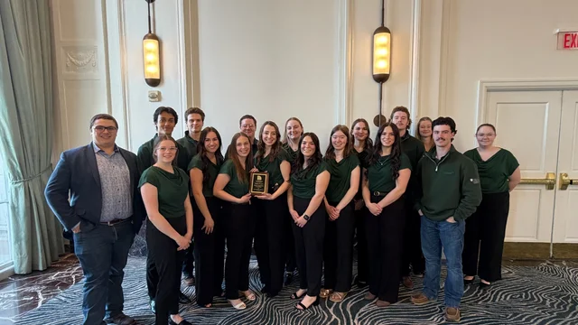 A group of fifteen young adults, most wearing dark green shirts and black pants, pose for a photo in a formal room. One woman in the center holds a small plaque.